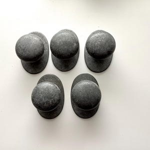 May include: Five dark grey cabinet knobs with a textured, weathered appearance. Each knob has a rounded top and an oval base. The knobs are arranged in a slightly offset pattern against a white background.