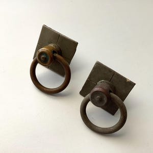 May include: Two antique bronze ring pulls with square backplates. The ring pulls are a dark bronze color, and the backplates have a textured, aged appearance. The hardware is likely for a cabinet or drawer.