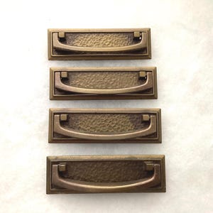 May include: Four rectangular brass drawer pulls arranged vertically. Each pull features a central hammered texture panel framed by a smooth border and a curved, hinged drop handle attached to two small square mounting points on the top edge.