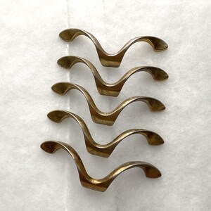 May include: A set of five brass-toned metal drawer pulls, each featuring a distinct wave-like, curved silhouette with a central mounting point and two outward-sweeping arms that taper at the ends, arranged in a vertical column on a white surface.