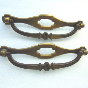 May include: Two matching brass drawer pulls featuring a horizontal backplate with a central rectangular cutout and a curved, swinging handle. The metal hardware has a muted, aged finish with rounded ends and a decorative center detail on the handle.