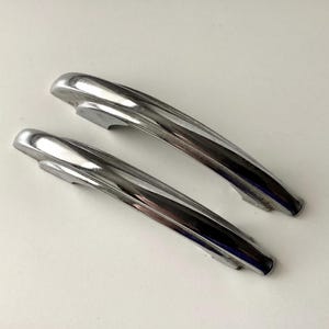 May include: Two vintage chrome door handles with a streamlined design. The handles are curved and have a shiny, reflective surface. They are likely from a classic car or a similar vehicle.