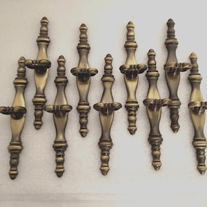 May include: A collection of ten antique-style cabinet pulls. Each pull is made of brass-colored metal with a detailed, ornate design. The pulls have a central decorative element and a handle on each side, perfect for furniture restoration.