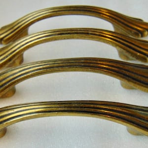 May include: Four antique-style cabinet handles with a curved, arched design. The handles are made of a brass-colored metal with a ridged texture and decorative ends. These handles are suitable for furniture restoration or adding a vintage touch.