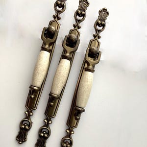 May include: Three antique-style cabinet pulls with a cream-colored, speckled center and ornate bronze-colored metal detailing. The pulls have a decorative design at each end and a central handle.