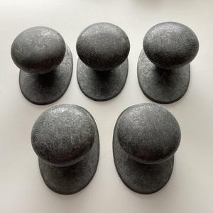May include: Five dark gray metal cabinet knobs with a textured finish. Each knob has a rounded top and an oval base. The knobs are arranged in a pattern on a white surface.