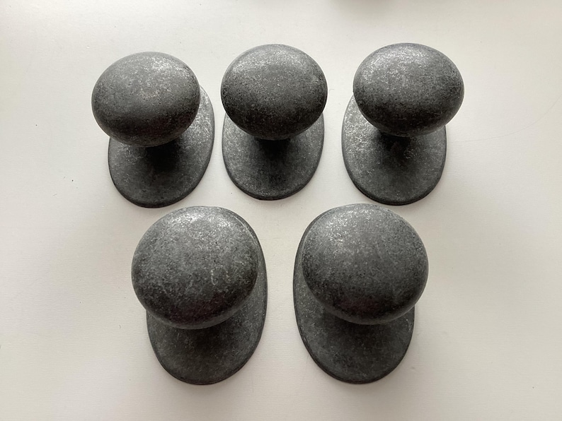 May include: Five dark grey metal cabinet knobs with a textured finish. Each knob has a rounded top and an oval base. The knobs are arranged in a pattern on a white surface.