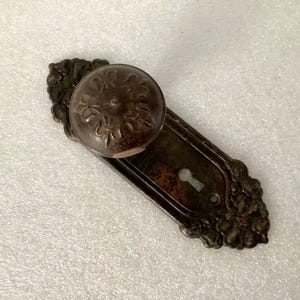 May include: An antique door knob and backplate set. The knob is a dark brown, round shape with a floral design. The backplate is rectangular with ornate edges and a keyhole. The metal has a tarnished, aged appearance.