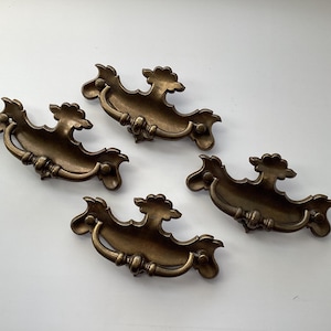 May include: Four antique brass drawer pulls with ornate, curved designs. Each pull features a decorative handle and a stylized floral or leaf motif. The hardware has a warm, aged patina, suggesting vintage or retro style.