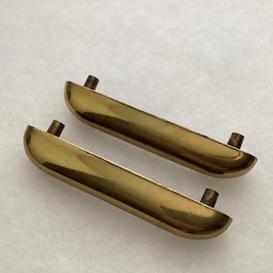 May include: Two vintage brass drawer pulls. Each pull is a curved, rectangular shape with a polished finish. The pulls have two small, cylindrical posts for mounting. The hardware is a warm, golden color.