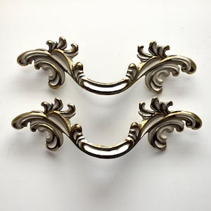 May include: Two ornate, vintage-style drawer pulls. Each pull features a curved, brass-toned handle with decorative, white and gold scrollwork on either end. The pulls are designed for furniture restoration or decorative projects.