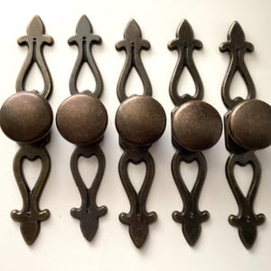 May include: Five antique bronze cabinet pulls with a decorative design. Each pull features a round knob and an ornate backplate with a heart-shaped cutout. The pulls are arranged in a row against a white background.