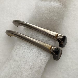 May include: Two vintage metal drawer pulls with a silver finish and dark, rectangular ends. The pulls are curved and have a small notch at the end. They are set against a white, textured background.