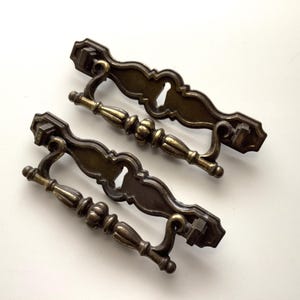 May include: Two antique brass drawer pulls with a decorative design. The pulls have a dark bronze finish with gold accents. Each pull has a keyhole and a handle with a detailed, ornate design. The pulls are approximately 6 inches long.