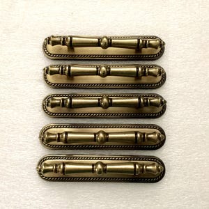 May include: Five antique brass cabinet pulls. Each pull features a bamboo-style design with a central rounded detail and decorative ends. The pulls are set on a rectangular backplate with a rope-like border. Hardware for furniture restoration.