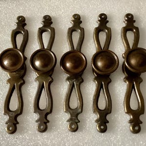 May include: Five antique bronze cabinet pulls. Each pull features a circular knob above an ornate, elongated handle with decorative cutouts. The pulls are arranged in a row, showcasing their aged, vintage appearance. Ideal for furniture restoration projects.