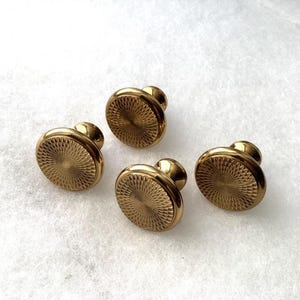 May include: A set of four round, gold-toned metal cabinet knobs. Each knob features a raised, textured sunburst pattern radiating from the center to the outer edge, with a smooth, polished rim and a short, cylindrical base for mounting.