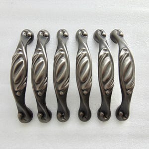 May include: Six antique bronze cabinet pulls with a decorative, flowing design. Each pull features a curved handle with an oval center and rounded ends. The pulls are arranged in a row, showcasing their ornate details and finish.