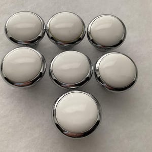 May include: Seven white and chrome cabinet knobs arranged in a triangular pattern. The knobs have a glossy white center and a shiny chrome base. The knobs are round and appear to be new.