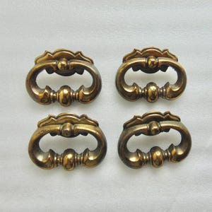 May include: Four antique brass drawer pulls with a decorative, ornate design. Each pull features a curved handle with a central, rounded detail and a scalloped top. The pulls have a warm, aged patina, suggesting vintage or retro style.