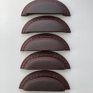 May include: Five dark brown, semi-circular cabinet pulls with decorative edges and a central recessed area. The pulls are arranged vertically against a white background, showcasing their ornate design and potential for furniture hardware.