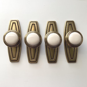 May include: Four vintage-style cabinet pulls. Each pull features a round, off-white ceramic knob set within a brass-toned metal frame. The frames have a rectangular shape with a decorative, geometric design. The pulls are arranged on a white surface.