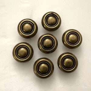 May include: Seven antique brass cabinet knobs. Each knob has a circular design with concentric rings and a raised center. The knobs have a vintage, aged appearance, suggesting they are old or have been treated to look old.