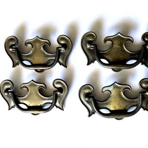 May include: Four antique brass drawer pulls with a decorative, ornate design. Each pull features a curved handle with scrolled ends and a central, stylized shield shape. The pulls are arranged on a white background.