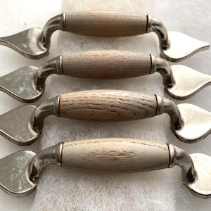 May include: Four cabinet pulls featuring a central light-toned wooden handle with a visible grain, connected to polished silver-colored metal ends shaped like stylized leaves or teardrops, arranged in a vertical stack on a textured surface.