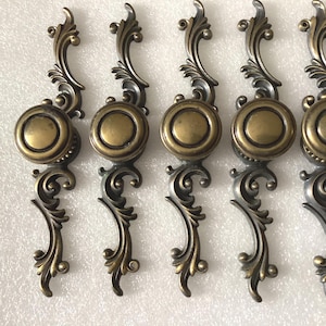 May include: Set of five antique brass cabinet knobs with ornate scrollwork and a round, slightly raised center. The knobs have a distressed finish and are perfect for adding a touch of vintage charm to any home.