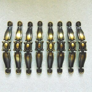 May include: Eight antique gold-toned cabinet drawer pulls with ornate designs. Each pull has a decorative oval in the center.