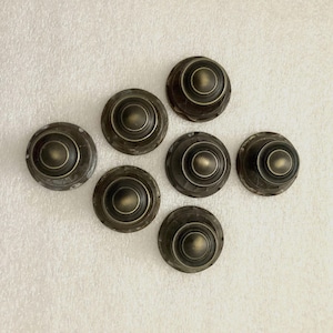 May include: Seven antique brass cabinet knobs arranged on a white surface. Each knob has a circular design with concentric rings and a central, rounded boss. The knobs have a weathered, aged appearance.