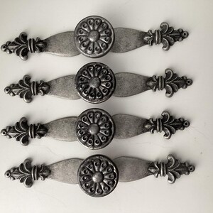 May include: Four antique-style cabinet pulls with a distressed silver finish. Each pull features a central circular knob with a floral design, flanked by decorative, stylized flourishes on a curved base. These hardware pieces are ideal for furniture restoration.