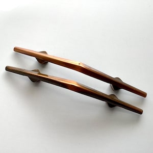 May include: Two bronze-colored cabinet pulls with a geometric design. The pulls are long and slender, with a slightly angled shape and a central mounting point. The finish has a warm, metallic sheen, suggesting a vintage or antique style.