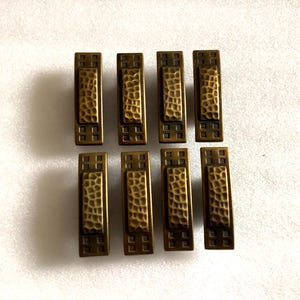 May include: Eight antique brass cabinet pulls with a hammered finish and a square design. The pulls have a rectangular shape with a raised center section.