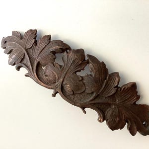 May include: A detailed, antique-style, brown leaf carving. The decorative piece features multiple leaves with intricate veining and a slightly curved shape. The carving is likely made of wood and has a weathered appearance, suggesting age and use.