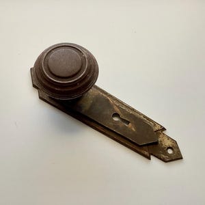 May include: Antique bronze door knob and backplate. The knob is round with concentric circles, and the backplate has a keyhole and decorative edges. The hardware shows signs of age and wear.