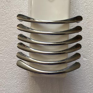 May include: A set of six curved, polished chrome cabinet pulls arranged vertically on a white rectangular backing. The pulls have a smooth, reflective surface, and are evenly spaced. The background is a textured white surface.