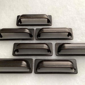 May include: Seven dark bronze metal drawer pulls arranged in a pyramid formation. Each handle features a rectangular backplate with rounded corners and a curved, arched grip section, showcasing a smooth, uniform finish across all hardware pieces.