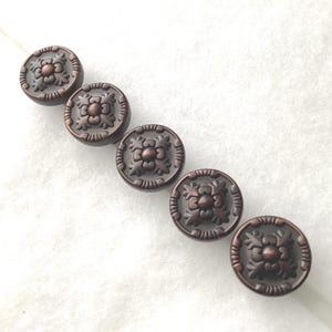 May include: Five circular buttons arranged in a diagonal line. Each button features a dark copper-toned finish with a raised central floral motif surrounded by a textured border. The design includes symmetrical leaf-like patterns radiating from the center.