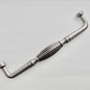 May include: A silver-toned cabinet pull with a decorative, ribbed center section and curved ends. The hardware is designed for easy installation and adds a touch of elegance to furniture. The pull is approximately 6 inches long.