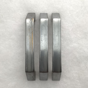 May include: Three vertical silver-toned metal drawer pulls arranged side by side. Each handle features a smooth, slightly arched rectangular design with a metallic finish. The leftmost handle shows minor signs of wear with a faint orange discoloration.