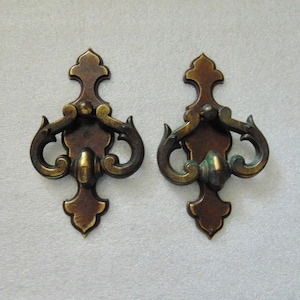 May include: Two ornate brass cabinet pulls with a decorative, scrolled design. The pulls are in a dark brass finish with a slightly aged patina.