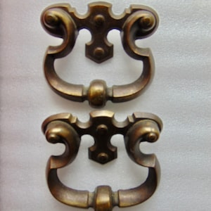 May include: Two matching antique-style brass drawer pulls featuring a decorative backplate with a central vertical post and two symmetrical scrolling arms that curve downward to meet a central rounded handle grip with a smooth metallic finish.