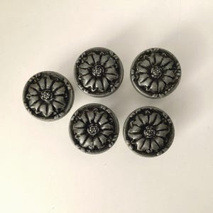 May include: Five antique-style cabinet knobs with a floral design. The knobs are circular, with a raised flower pattern in the center and a detailed border. The finish is a muted silver color, giving them a vintage appearance.