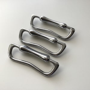 May include: Four shiny, silver-colored metal handles are arranged diagonally on a white surface. Each handle has a curved, elongated shape with a central, rounded section. The handles appear to be new and unused.