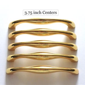 May include: Five gold-colored cabinet pulls with a curved design. The pulls are 3.75 inches (9.5 cm) center to center, as indicated by the text above the pulls. The pulls have a shiny, reflective surface.