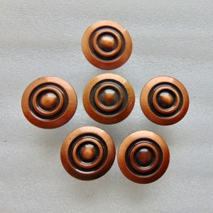 May include: Seven antique-style copper-colored cabinet knobs arranged in a circular pattern. Each knob features a central raised circle surrounded by concentric rings, creating a classic, decorative design. The knobs have a warm, metallic finish.