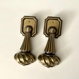 May include: Two antique brass drawer pulls. Each pull features a rectangular backplate, a fluted center, and a decorative, spiral-shaped bottom. The hardware is a warm, aged brass color, suggesting vintage charm and a classic aesthetic.