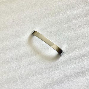 May include: A shiny, silver-colored cabinet pull handle with a curved design. The handle is rectangular with rounded edges and a polished finish, reflecting light. It is likely made of metal and designed for furniture.